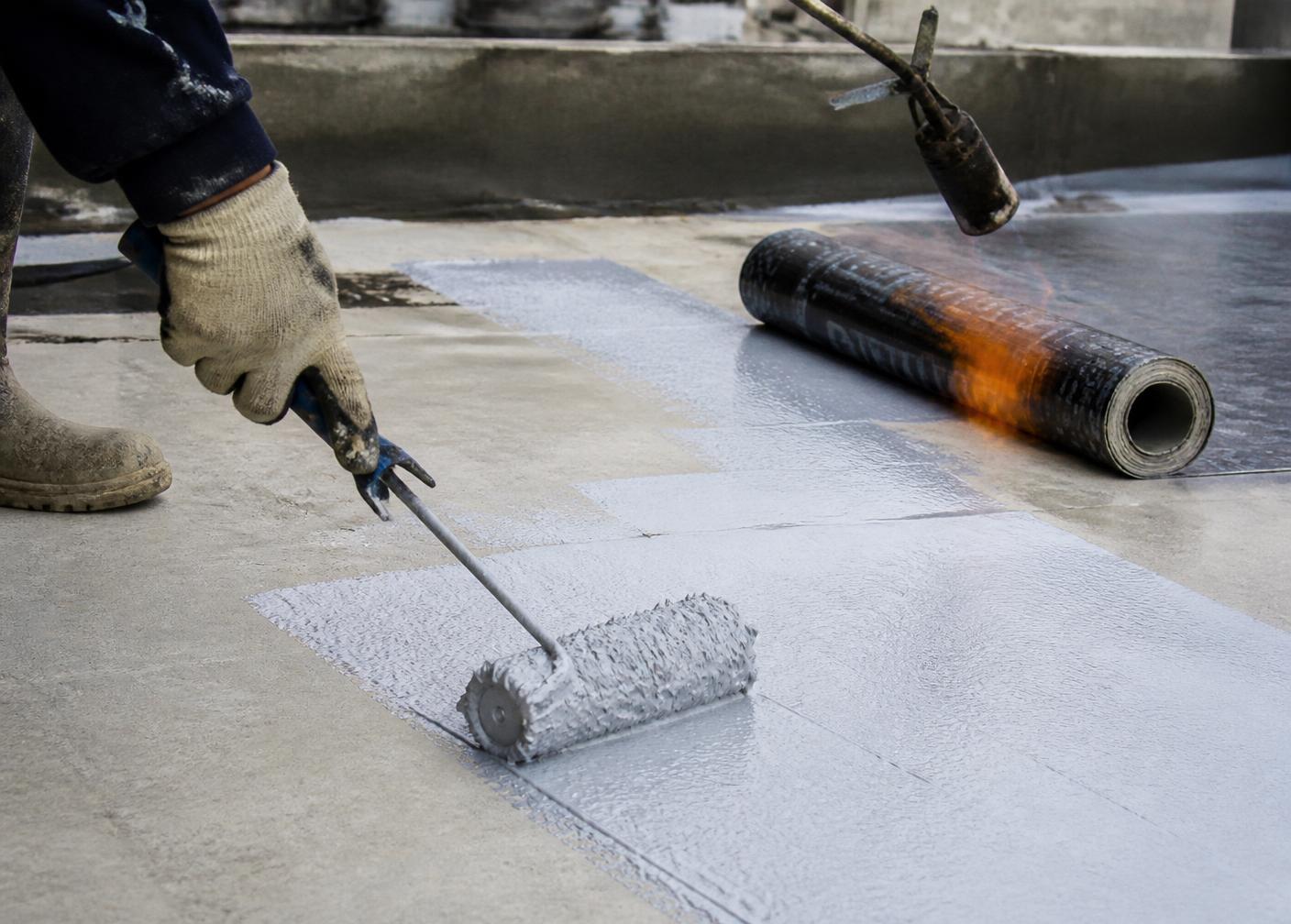 Applicator rolling liquid waterproofing coating on a concrete terrace beside a bitumen membrane roll